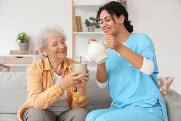 Young African-American female medical worker pouring elderly woman cup of tea in nursing home
