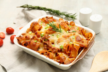 Baking dish with tasty pasta, rosemary, cut green onion and tomatoes on light grunge background