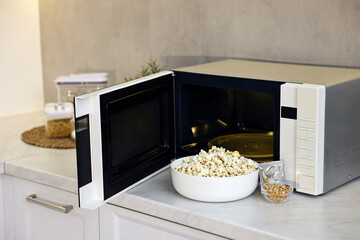 Open microwave oven with bowl of popcorn, kernels and bag on white marble countertop in kitchen