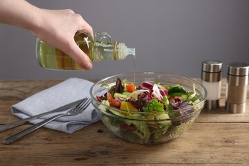 Woman pouring oil onto salad at wooden table against grey background, closeup