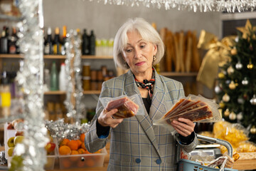 While shopping on eve of New Year holidays, elderly lady is interested in packing hamon. She has collected basket of products and chooses hamon, examines vacuum-packed meat delicacy.