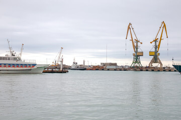 Industrial port scene with ships docked near large yellow cranes under a cloudy sky. Calm water reflects the vessels and equipment, showcasing a busy harbor with cargo and maritime activity