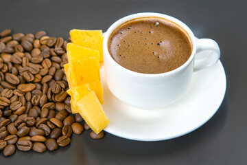 Roasted coffee beans surround a white cup of espresso and yellow cheese sticks on a dark surface, creating a warm, inviting composition.