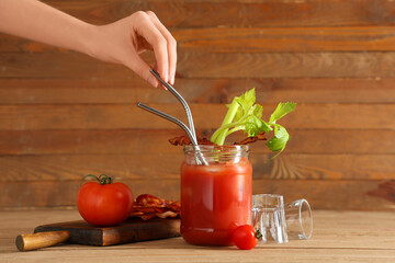 Female hand with straw and mason jar of bloody mary cocktail with bacon on wooden background