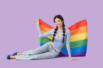 Beautiful young woman with rainbow flag sitting on lilac background. LGBT concept
