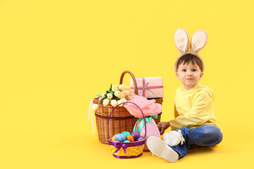 Cute little boy in bunny ears with Easter gifts sitting on yellow background