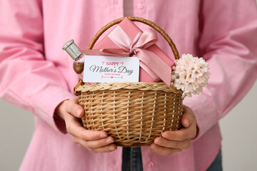 Young woman holding basket with flower, gift box and perfume for Mother's Day on white background