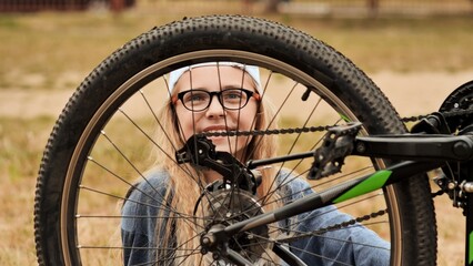 Teenage girl fixing bicycle wheel on grassy lawn, smiling while working on bike during sunny day outdoors