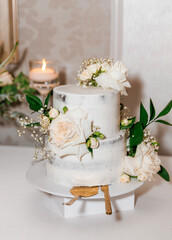 A white wedding cake decorated with white roses stands on the table at the banquet. A small two-tier holiday cake with vanilla cream decorated with fresh flowers. Sweet treats for guests
