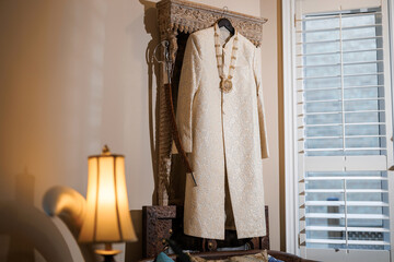Indian traditional outfit of the groom hangs on a hanger in the room. A festive wedding sherwani with a pattern and a necklace hangs above the bed. Prepared Indian wedding men's wear and Talwar saber