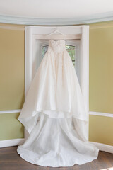 White wedding dress hanging on a hanger near the window. Beautiful designer wedding dress for the bride. Preparing for the wedding ceremony. Traditional dress for the wedding
