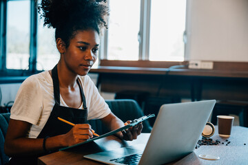 Black woman barista in her own coffee shop smiles confidently at counter with laptop. Self-employed entrepreneur manages business finance bookkeeping. Portrait of determined owner.