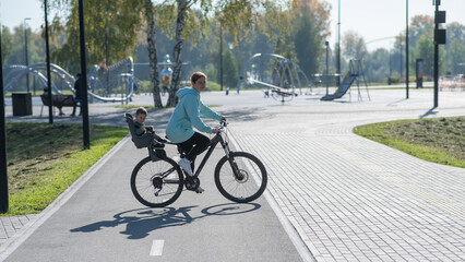 Fototapeta premium Caucasian woman riding bicycle with toddler son sitting behind her in child seat in park. 