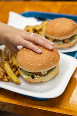 Hands grasp a juicy burger above golden fries, capturing the moment before indulgence in a casual, mouthwatering scene.

