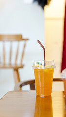 A refreshing orange drink in a clear cup with a straw, placed on a wooden table, with a blurred background of a cozy cafe setting.