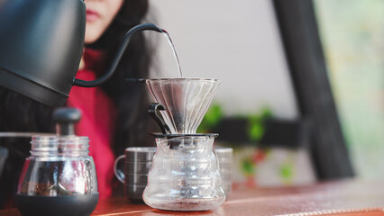 A person is brewing coffee using a pour-over method with a kettle, dripper, and glass carafe, set against a bright, cozy background.