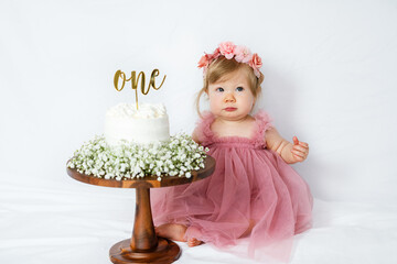Fototapeta premium Adorable baby girl's first birthday cake smash moment in a dreamy pink tulle dress with a flower crown. She sits beside a white frosted cake topped with 