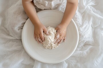 A child's hands gently knead a mound of white clay in a ceramic bowl.