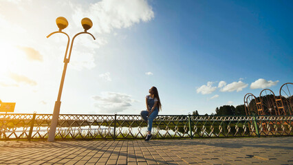 Serene woman relaxing on lakeside fence, basking in golden sunset light, absorbing peaceful natural landscape