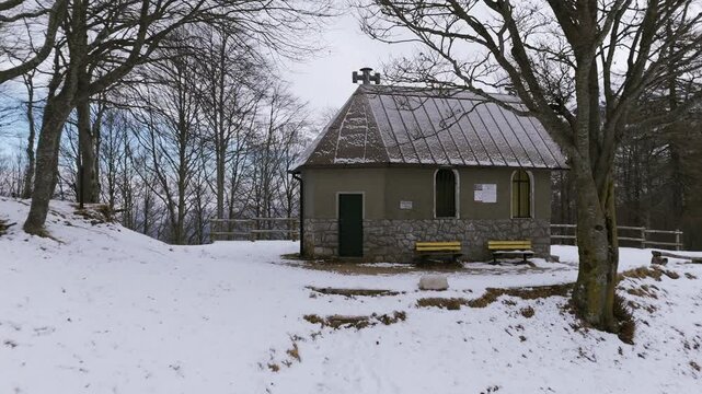 Hiker walking in snow with poles towards small mountain shelter in winter, 89th Brigade Bivouac near Bocchetta di Prada, Italy. Aerial drone at low altitude