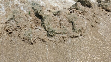Turbulent waves surge over sandy shore, mixing foam and sediment. The water is murky, revealing...