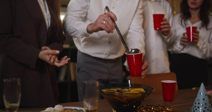 Close-up of a happy guy in a white shirt pouring punch into glasses during a corporate party in the office