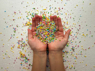 Child's hands are covered with multi-colored sugar sprinkles and form a heart shape. Child plays with small sprinkles topping.