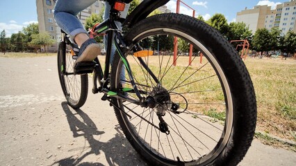 Low angle view of rear bicycle wheel with school girl riding bicycle in urban park during summer day