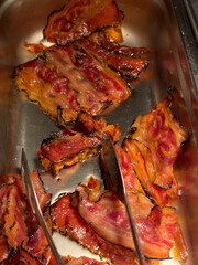 Crispy cooked bacon slices in a stainless steel tray, served warm with tongs at a buffet or breakfast station.