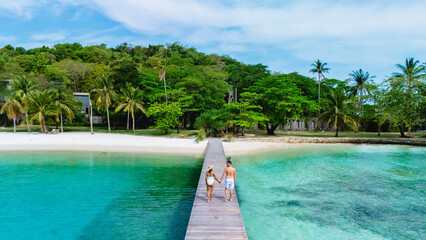 A romantic couple walks hand in hand along a wooden pier, surrounded by crystal-clear waters and lush greenery. The serene atmosphere captures the essence of paradise on Koh Kham Island Thailand