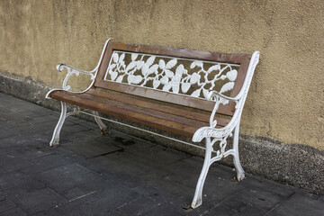 A rustic wooden garden bench with white cast iron ornate details, set against a textured beige wall and gray paved walkway. The bench features a decorative floral pattern on the backrest.