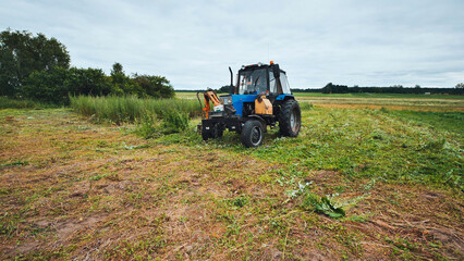 Agricultural tractor cutting grass with rotary mower, creating linear swaths on overcast farmland landscape