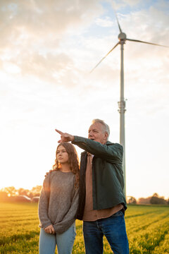 Grandfather and granddaughter in a field with a wind turbine at sunset