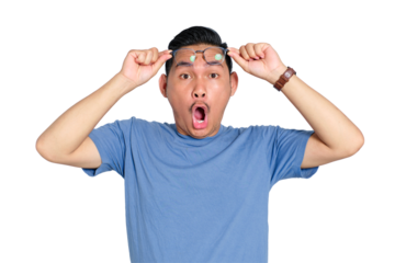 Surprised young Asian man in glasses holding eyewear with open mouth expression, wearing blue t-shirt, isolated on transparent background