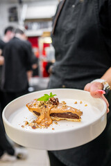 A professional chef presents a plated gourmet chocolate tart drizzled with caramel sauce, garnished with crushed nuts and a fresh mint leaf, in a busy restaurant kitchen.
