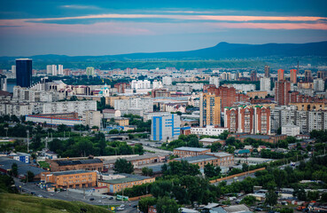 Obraz premium Krasnoyarsk cityscape revealing urban landscape, residential zones, winding streets, verdant greenery, mountain silhouettes against dramatic sunset lighting