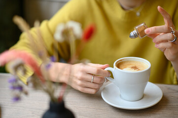 A woman’s hands sprinkling cinnamon into a cup of coffee at a cozy café. A warm, inviting scene capturing the essence of comfort, relaxation, and the simple pleasures of a coffee break.