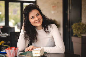 Happy smiling brunette curly woman sitting in cafe, ordering krempita cream cake. Positive emotions...