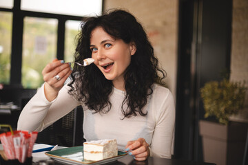 Young curly brunette woman eating cake with cream using fork sitting in cafe. Cheat meal day...