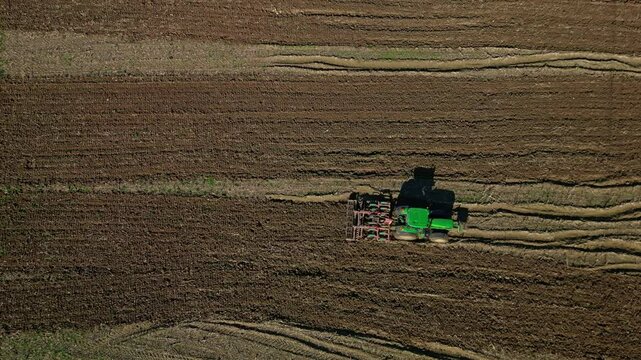 Top-down drone view of green tractor plowing brown farmland with parallel lines.
