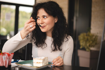 Young curly brunette woman eating cake with cream using fork sitting in cafe. Cheat meal day concept. Woman is preparing with appetite to eat eclair. Enjoy bakery in restaurant. Wow emotions.