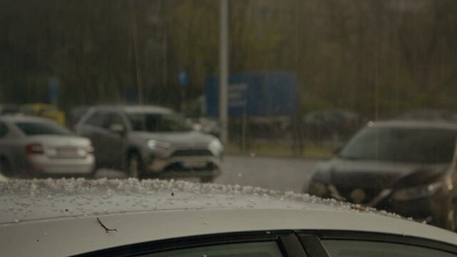 Hail pummels the car roof amidst heavy rain in a city, creating splashes of ice. The storm leads to potential damage and adds to the chaotic weather conditions observed.