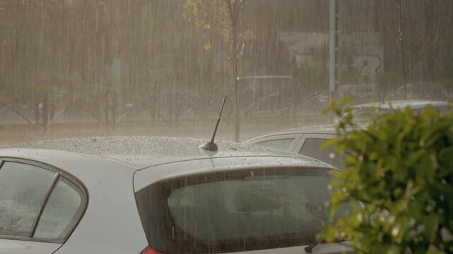 Hail and heavy rain batter the roof of a parked car in a city. The powerful storm produces splashes and possible damage due to the icy precipitation.