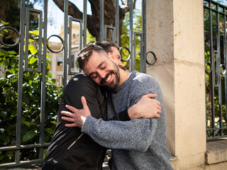 Two adult brothers enjoy a warm embrace in a sunny park. They stand beside a decorative fence...