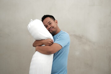 A Southeast Asian man in a light blue t-shirt, hugging a white pillow with a peaceful expression,...