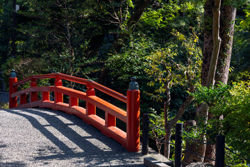 Traditional red wooden bridge at Tsurugaoka Hachimangu Shrine in Kamakura Japan casting shadows on a gravel path surrounded by lush green trees