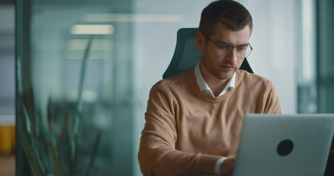 Smart looking focused businessman working on a laptop and thinking. Millennial managing family budget, handling financial paperwork, and tracking expenditures on a laptop at a modern open space office
