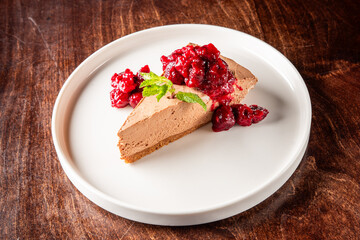 A slice of creamy chocolate cheesecake topped with vibrant berry compote and fresh mint leaves, elegantly presented on a round white plate against a rustic wooden background.