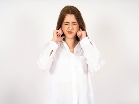 Caucasian young girl wearing white shirt  covering ears with fingers with annoyed expression for the noise of loud music. Deaf concept.