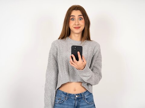 Caucasian young girl wearing grey sweater holds mobile phone in hands and rejoices positive news, uses modern cellular